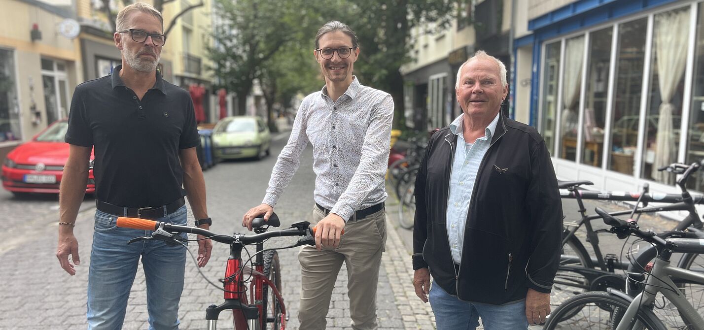 Drei Männer stehen auf einer Straße, jener in der Mitte hält strahlend ein Fahrrad am Lenker fest