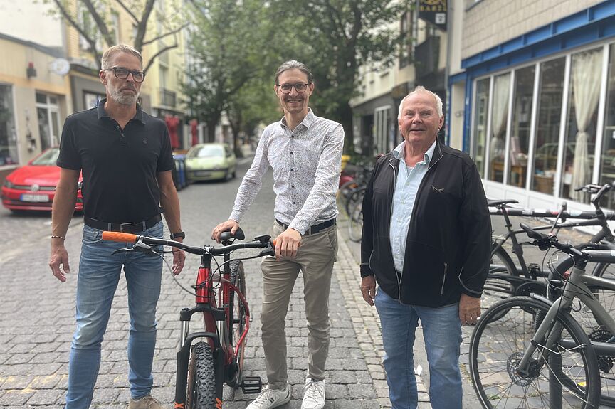 Drei Männer stehen auf einer Straße, jener in der Mitte hält strahlend ein Fahrrad am Lenker fest