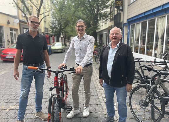 Drei Männer stehen auf einer Straße, jener in der Mitte hält strahlend ein Fahrrad am Lenker fest