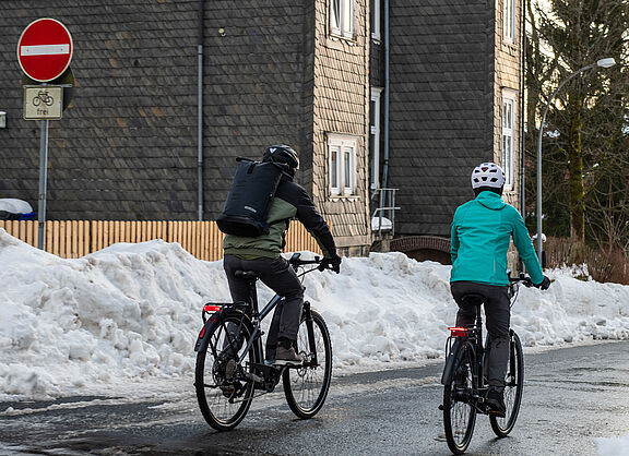 Zwei Fahrradfahrende in Winterjacken auf winterlicher Straße mit geräumtem Schnee an der Seite