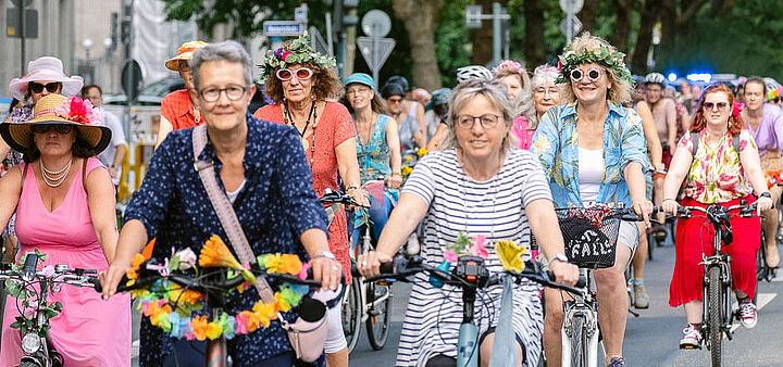 Bunt gekleidete Frauen auf geschmückten Fahrrädern lachend auf einer Straße radelndf ei