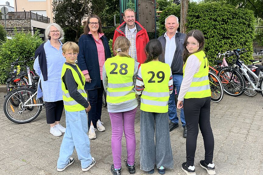 Mehrere Personen vor einer Schule stehend davon 3 Schüler in gelben Westen mit Rückennummernmit dem Rücken zu