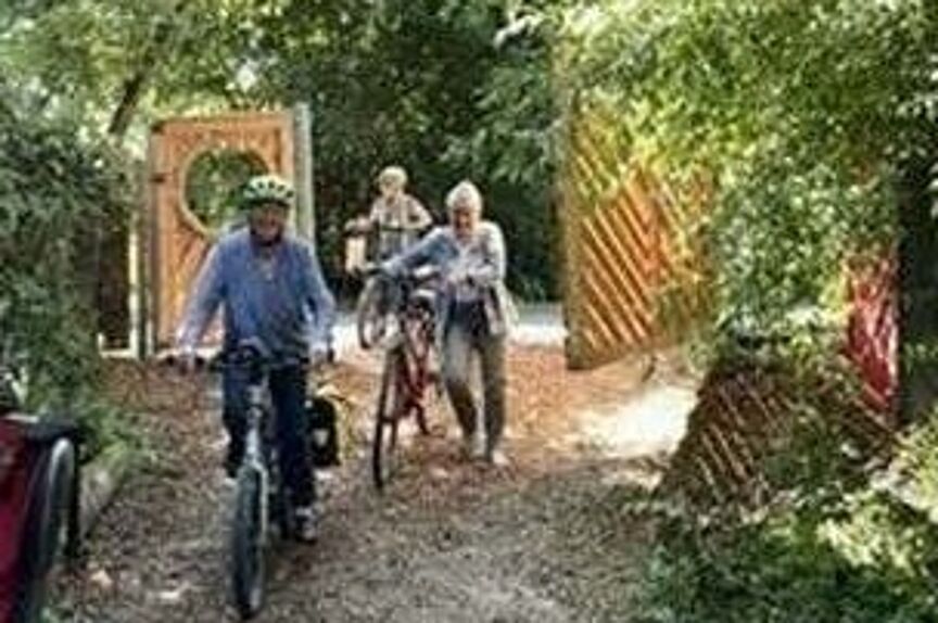 Drei sich auf einem Waldweg befindende Fahradfahrende . hinter ihnen ein offenes Holztor