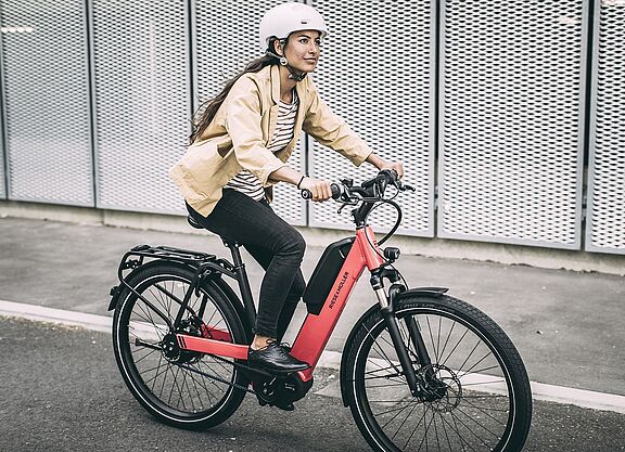 Elektroradtypen Eine Frau fährt auf einem roten Elektrorad auf Asphalt, im Hintergrund graue, gelochte Metallwände.